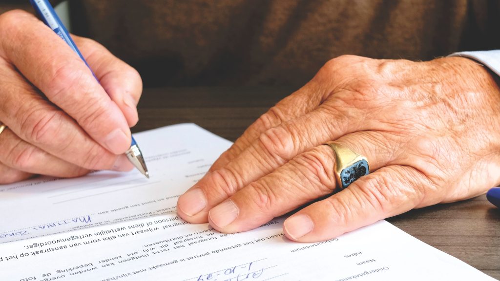 Close-up of a senior adult signing a legal document with a focus on hand and gold ring. Gros plan d'un adulte senior signant un document juridique avec un focus sur la main et la bague en or.