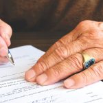 Close-up of a senior adult signing a legal document with a focus on hand and gold ring. Gros plan d'un adulte senior signant un document juridique avec un focus sur la main et la bague en or.